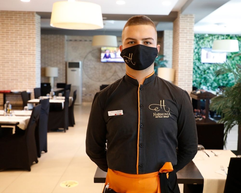 Ein Hotelangestellter steht im Restaurant. Er trägt einen Mund-Nasen-Schutz und schaut in die Kamera.