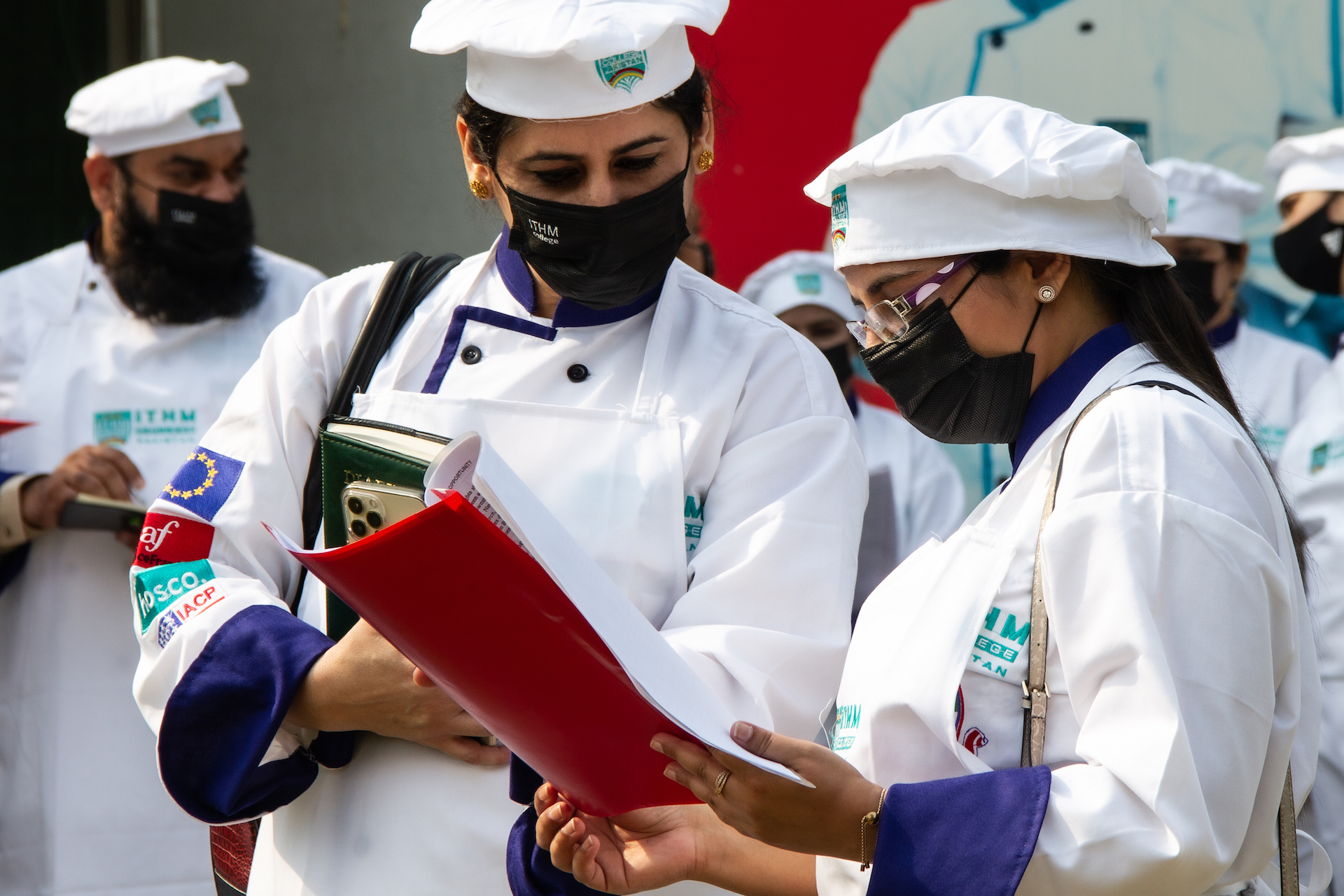 Zwei Frauen, die Kochjacken, Kochmützen und Mund-Nasen-Schutz tragen, schauen gemeinsam in einen Hefter mit Unterlagen. 