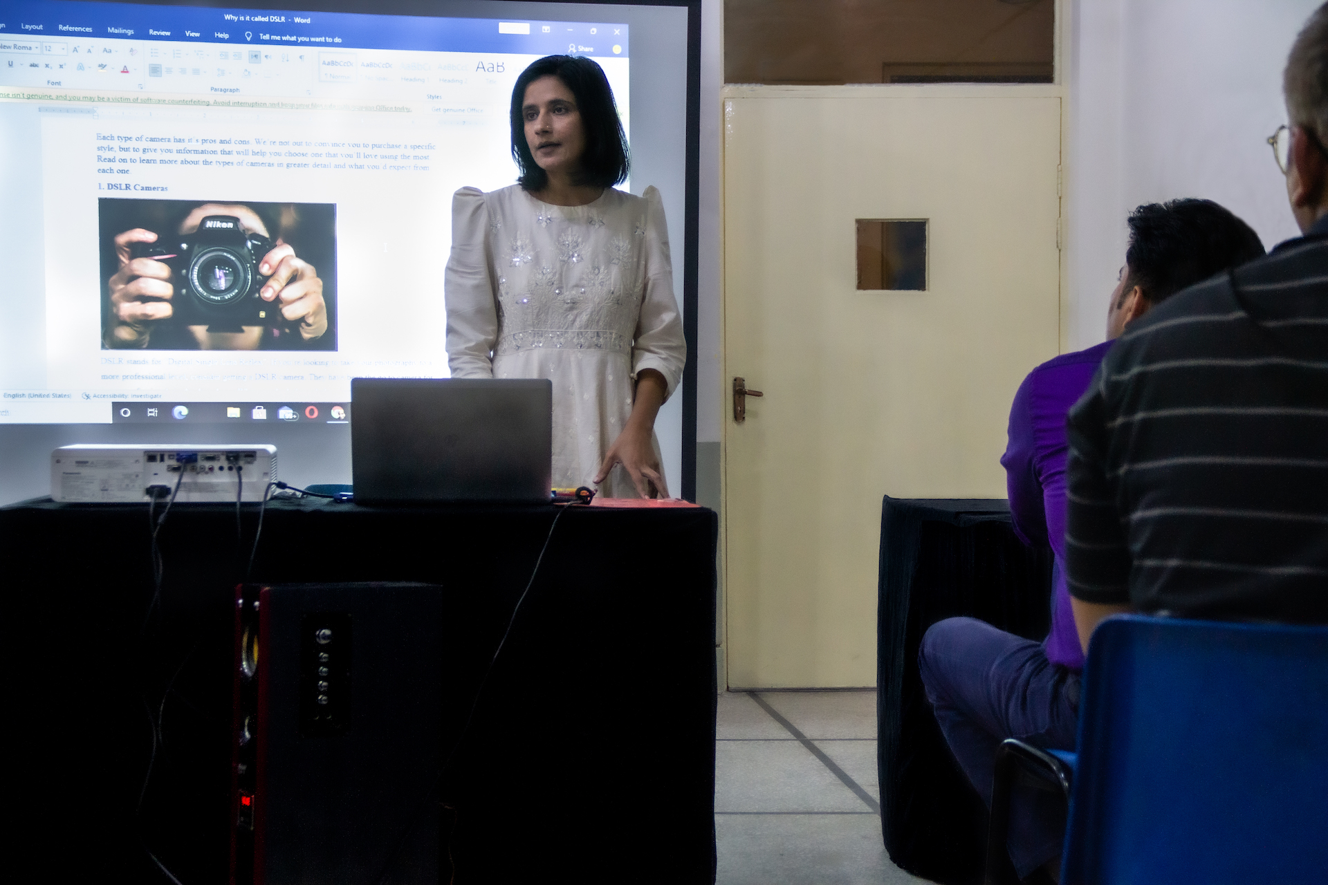 Eine junge Frau mit mittellangem, schwarzem Haar steht in einem Klassenraum vor einer Klasse. Ein Beamer wirft eine Präsentation hinter sie an die Wand, in der es um Kameras geht. 