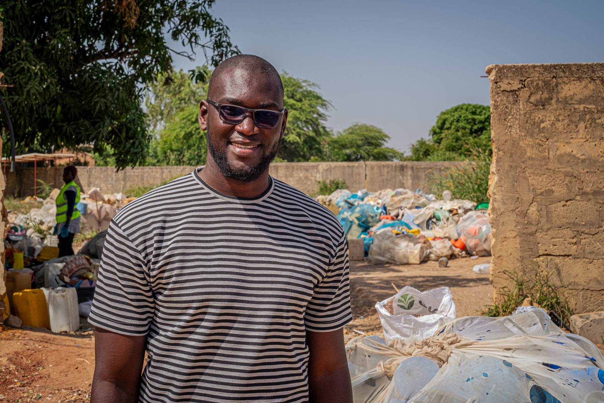 Ein Mann mit gestreiftem T-Shirt und Sonnenbrille steht vor einem Platz voller gefüllter Müllsäcke. Er lächelt in die Kamera. 