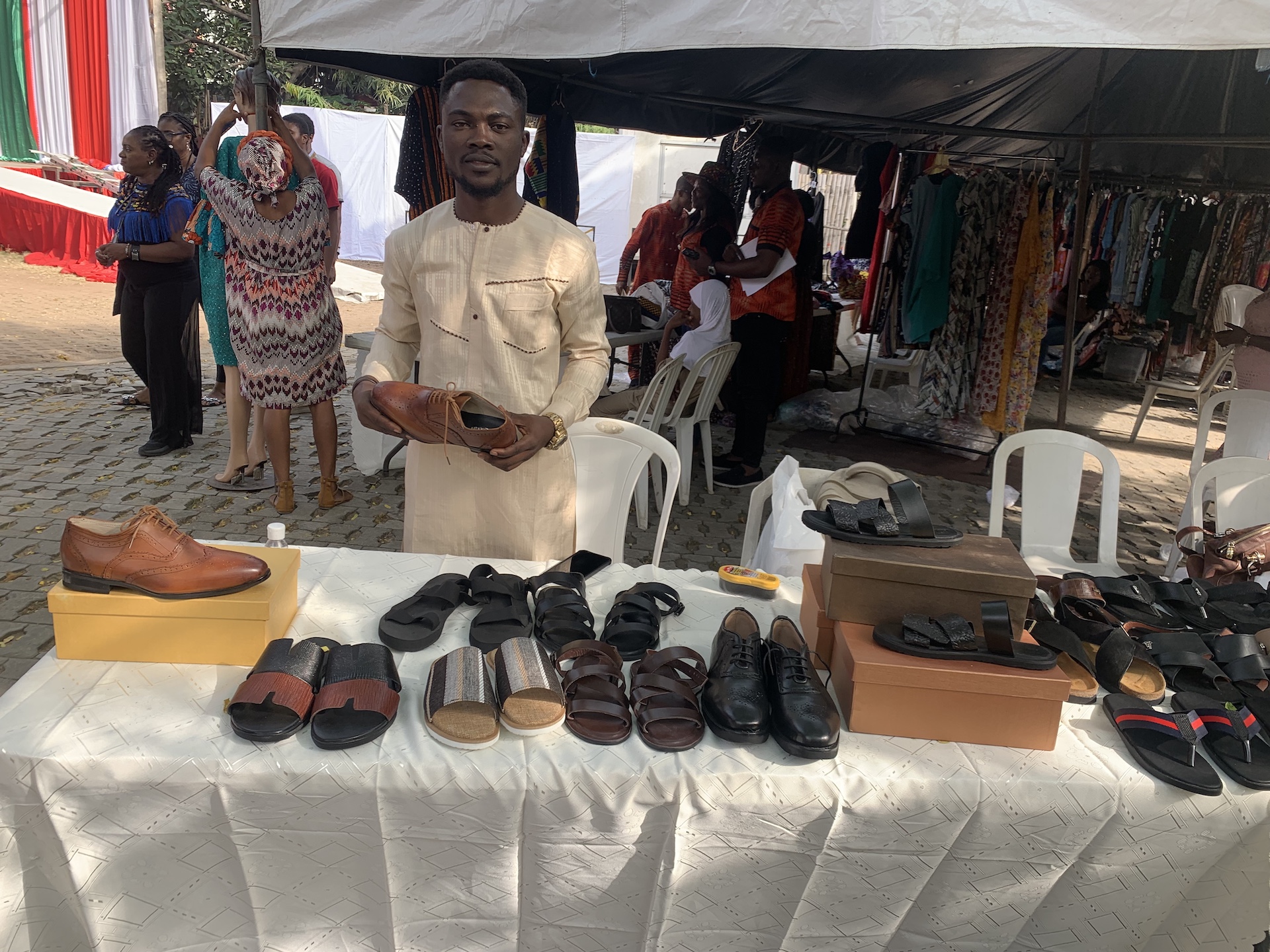 Ein Mann in einem beigefarbenen Oberteil steht hinter einem Marktstand und schaut in die Kamera. Vor ihm auf der Auslage stehen Freizeit- und Büroschuhe für Herren in Schwarz und Braun. Er hält einen braunen Herrenschuh in der Hand.
