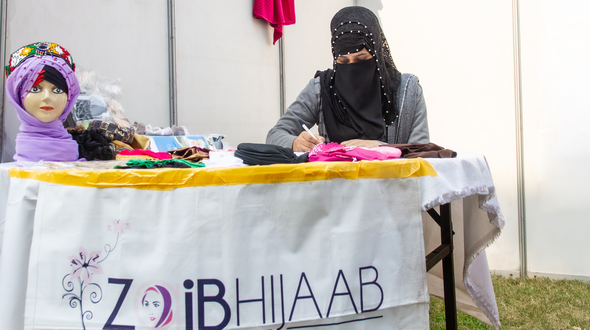 Eine Frau sitzt an einem Tisch. Sie hält einen Stift in der Hand, mit dem sie etwas schreibt. Auf dem Tisch liegen Hijabs in verschiedenen Farben. Auf einem Schild steht der Name ihres Unternehmens: „Zaib Hijaab“.