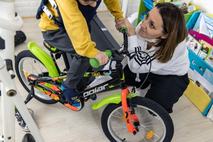 Ein kleiner Junge sitzt auf einem Kinderfahrrad mit beiden Füßen auf den Pedalen. Eine Frau hockt daneben und stützt den Jungen.