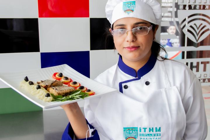 Eine Frau mit Kochjacke und Kochmütze balanciert einen Teller mit einem schön angerichteten Fischgericht auf ihrer rechten Hand und sieht in die Kamera. 