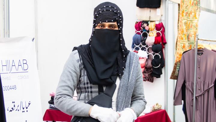 Eine Frau mit schwarzer Nikab steht vor einer weißen Wand. Man sieht nur ihren Oberkörper, sie trägt eine hellgraue Jacke und weiße Handschuhe. Rechts im Hintergrund steht eine Kleiderstange, an der Abayas hängen.
