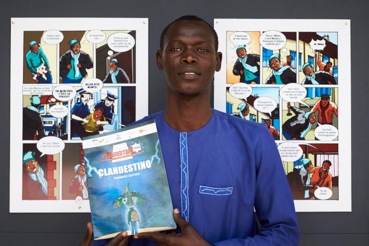 Ein junger Mann in marineblauer Bekleidung steht vor einer dunkelgrauen Wand und schaut in die Kamera. In den Händen hält er den Comic mit dem Titel „Clandestino“. An der Wand hinter ihm hängen zwei Seiten aus dem Comic in großem Format.