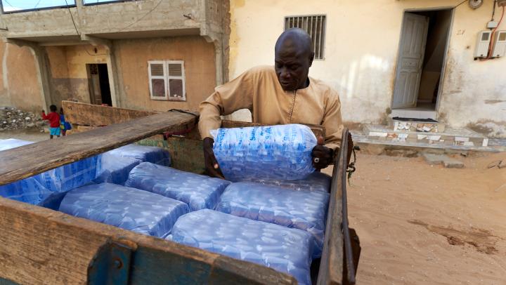 Ein Mann in einem hellbraunen Oberteil legt eine große Plastiktüte, die mehrere kleine Plastiktüten mit Wasser enthält, zu vielen anderen Tüten auf einen Holzwagen. 