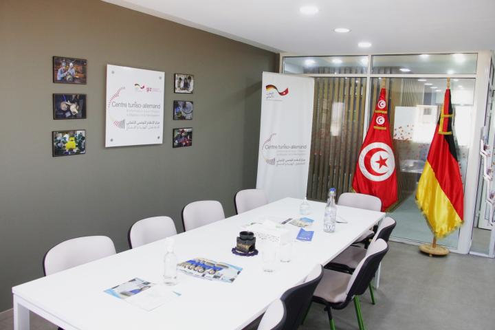 In einem Raum mit dunkelgrauem Boden steht ein weißer, langer Tisch mit neun Stühlen. An einem Ende des Tischs stehen vor einer Glaswand die tunesische und die deutsche Flagge. Ein Schild an der Wand und ein Rollup-Banner tragen den Zentrumsnamen. 