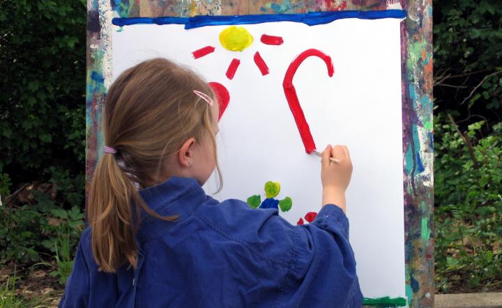 Ein Mädchen mit einem Zopf ist von hinten zu sehen, sie malt mit einem Pinsel ein Bild auf eine Leinwand. Im Hintergrund sind Büsche zu sehen. 