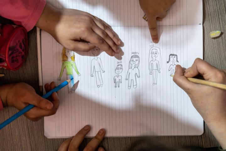 Eine Kinderzeichnung in einem Schulheft zeigt eine Familie. Rund um das Bild sind Kinderhände mit Stiften zu sehen.