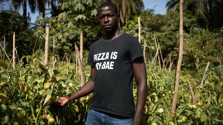 Ein junger Mann mit kurzen Haaren trägt ein schwarzes T-Shirt und Jeans. Er steht vor einem Feld mit Pflanzen und Palmen und schaut in die Kamera.