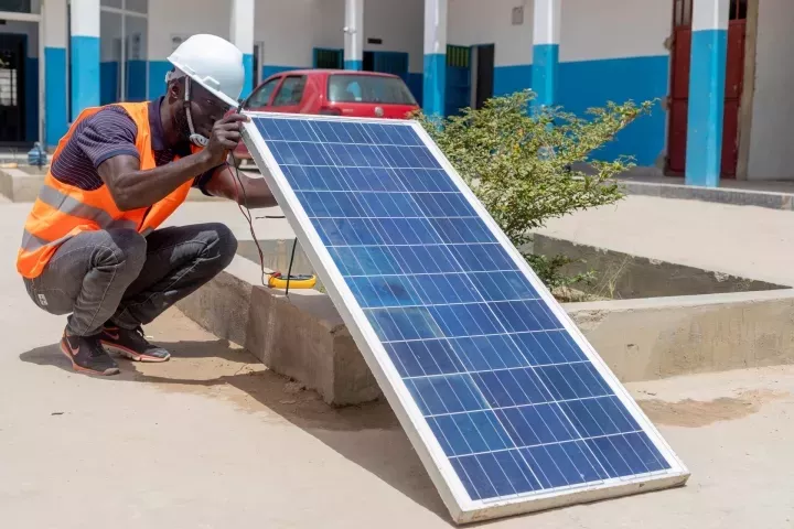  Ein junger Mann in dunkelblauem T-Shirt, orangefarbener Warnweste und weißem Helm hockt in einem Innenhof. Er hält ein Solarpanel in der Hand, dessen Rückseite er begutachtet.  