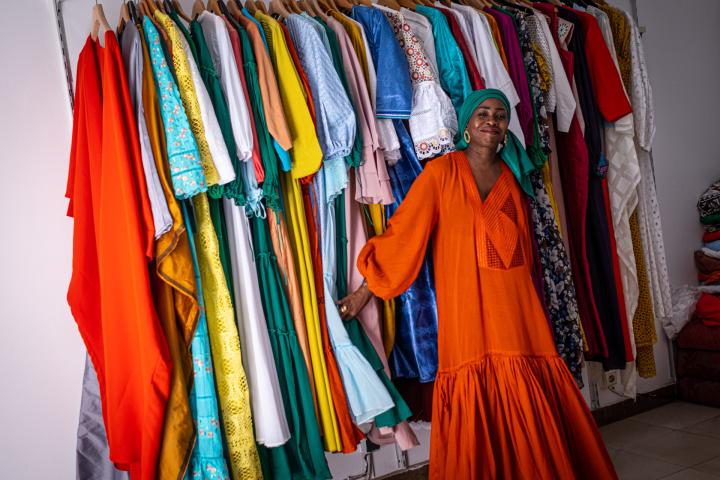 Eine Frau in langem, leuchtend orangefarbenem Kleid steht selbstbewusst vor einer Kleiderstange, an der viele bunte Kleider hängen.