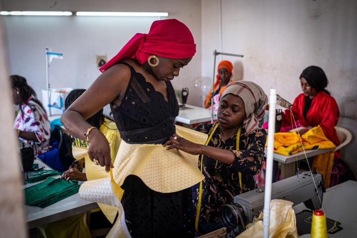 In einer Schneiderwerkstatt sitzen Frauen an Nähmaschinen. Bei einer Frau im Vordergrund wird Maß für eine Kleid genommen. 