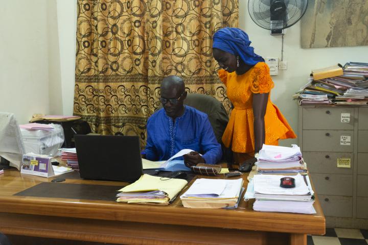 Ein Mann sitzt an einem Schreibtisch mit Akten und einem Laptop. Eine Frau in orangefarbenem Kleid steht daneben und blickt ihm über die Schulter. 