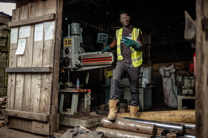 Ein junger Mann steht im Eingang einer Werkstatt mit Maschinen und lächelt.