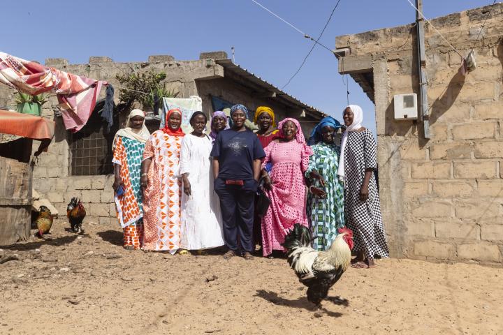 Eine Gruppe von Frauen steht zwischen Gebäuden, vor und neben ihnen laufen Hühner.