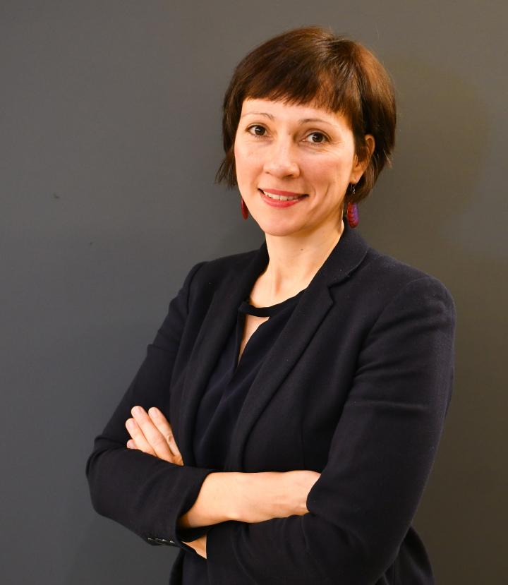 Eine Frau mit kurzem braunem Haar lächelt mit verschränkten Armen in die Kamera. Sie ist schwarz gekleidet.