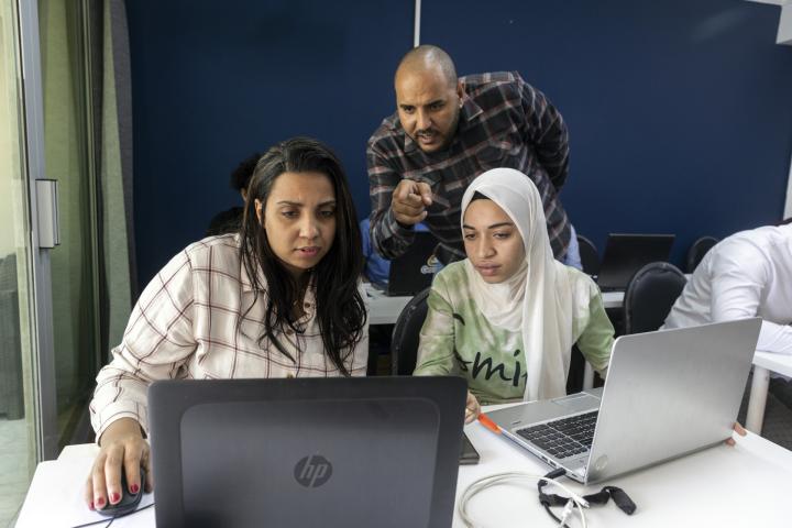  Zwei Frauen sitzen vor Laptops. Eine von ihnen trägt ein Kopftuch. Mitten zwischen ihnen steht ein Mann und zeigt auf einen der Bildschirme. 