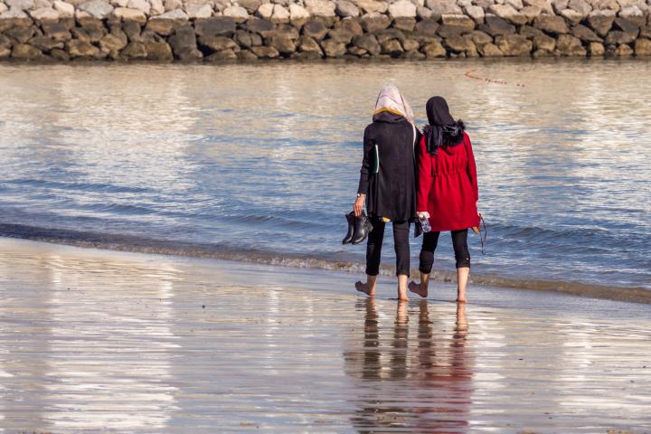 Zwei Frauen gehen am Strand entlang. Sie sind von hinten zu sehen und tragen Kopftücher. 