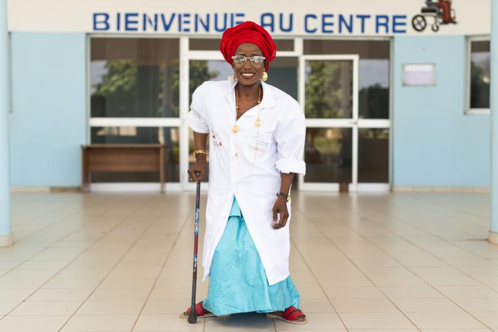 Eine Frau mit roter Kopfbedeckung schaut selbstbewusst in die Kamera. Sie stützt sich auf eine Gehhilfe und steht vor dem Rehabilitationszentrum, auf dem in Französisch „Willkommen im Zentrum“ steht. 