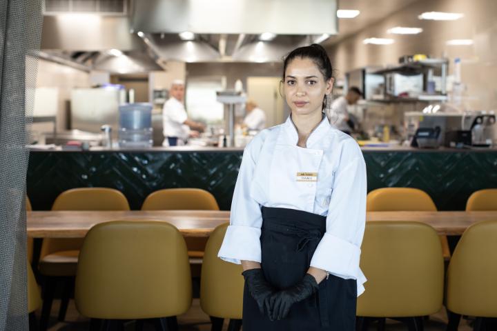 Eine junge Frau mit dunkelbraunen Haaren blickt in die Kamera. Sie trägt die Dienstkleidung einer Hotelküche. Im Hintergrund sieht man einen Küchenbetrieb. 
