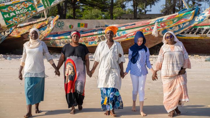 Fünf Frauen stehen am Strand und halten sich an den Händen. Sie blicken in die Kamera. Im Hintergrund sind bunt bemalte Fischerboote.