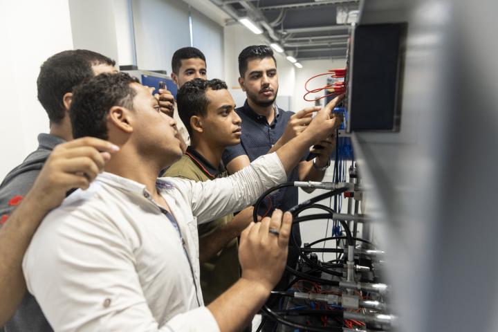 5 junge Männer stehen vor einer elektronischen Schaltzentrale und schauen konzentriert.