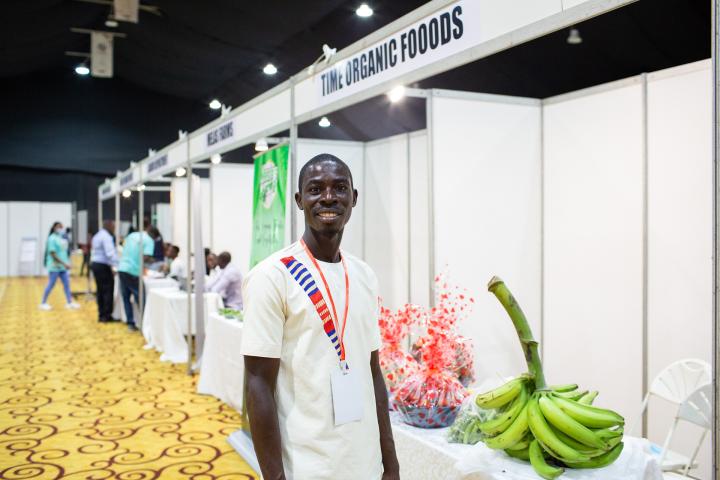 Ein junger Mann in weißem T-Shirt steht an einem Messestand in einer Halle. Rechts neben ihm eine Bananenstaude. 