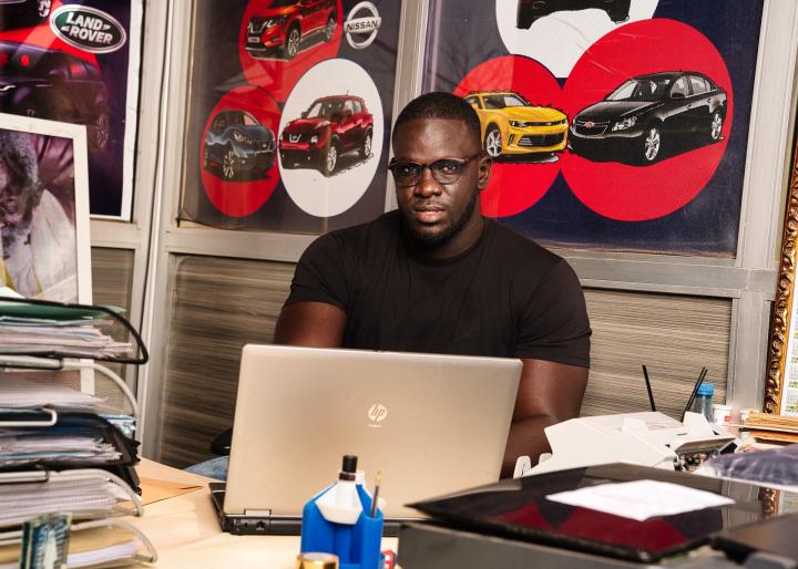  Ein Mann sitzt mit einem Laptop am Schreibtisch. Hinter ihm an der Wand Zeichnungen von Autos.  