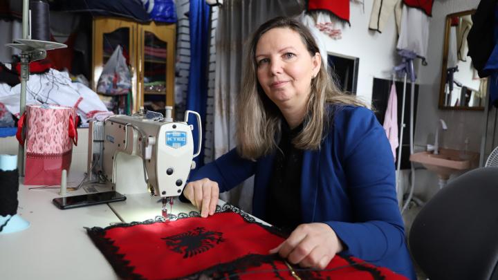  Eine Frau sitzt in einer Schneiderei an einer Nähmaschine und schaut freundlich in die Kamera.  